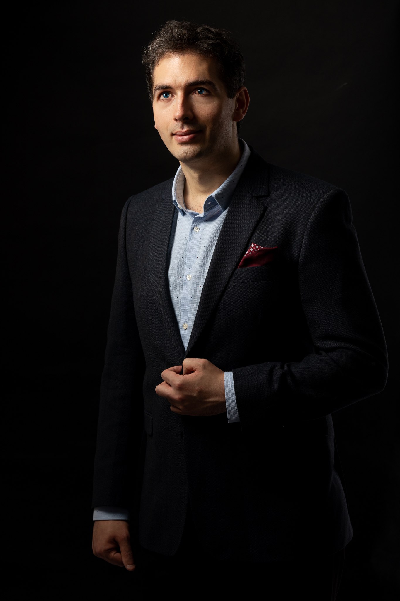 Formal portrait of Christian – tall man with brown hair and blue eyes wearing a suit, against a black background.