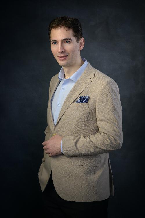 Formal portrait of Christian – tall man with brown hair and blue eyes wearing a suit, against a black background.